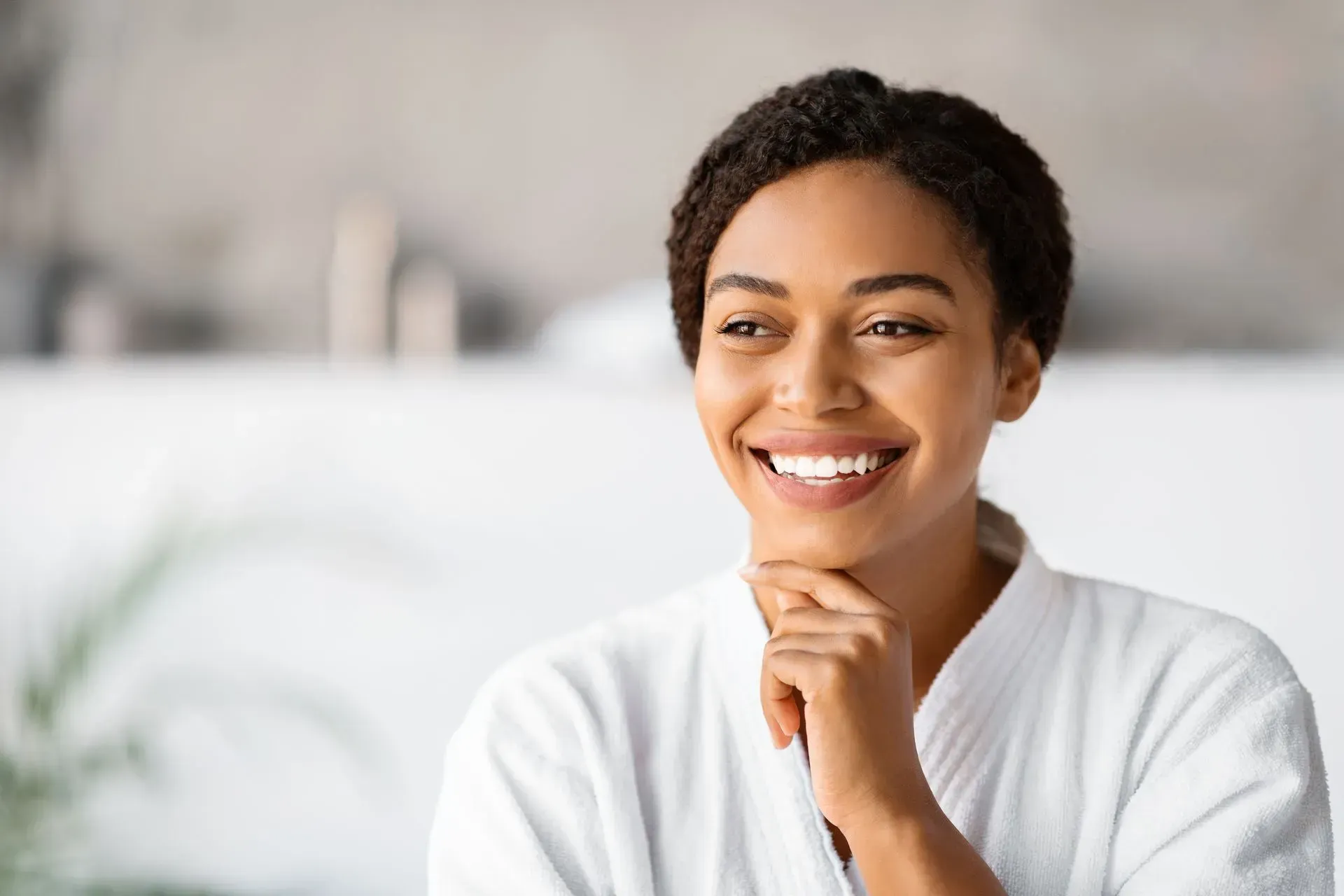 Smiling person in a white robe resting their chin on hand in a bright room