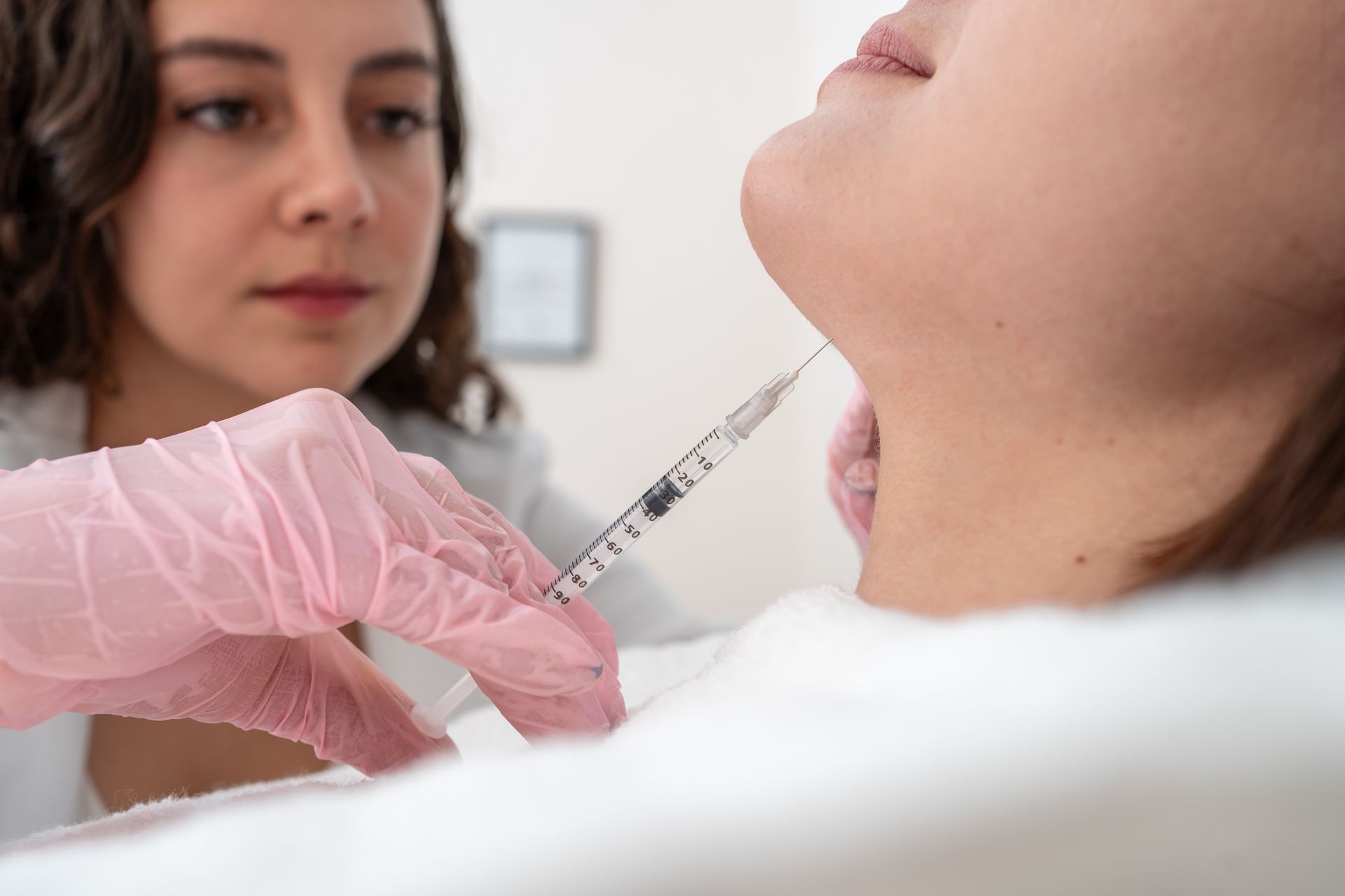 A gloved clinician injects the neck with a syringe while another person watches nearby.