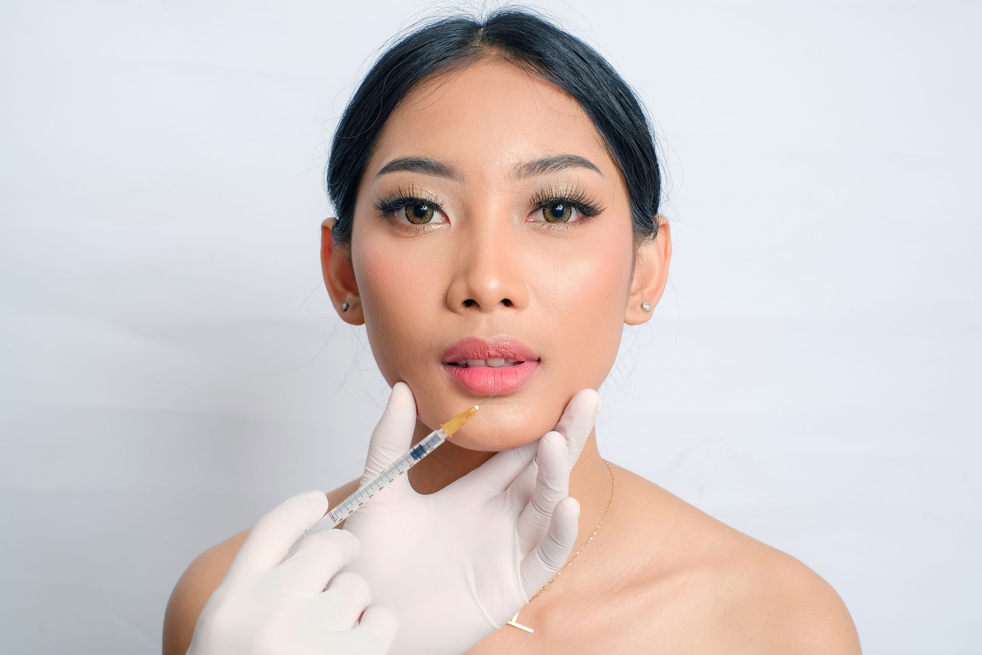 Cosmetic injector applying filler to a woman’s chin with a syringe, wearing white gloves.