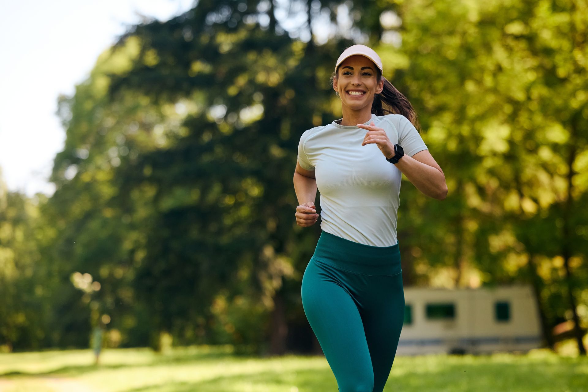 Woman running in a park, smiling in a white top and teal leggings at sunset.
