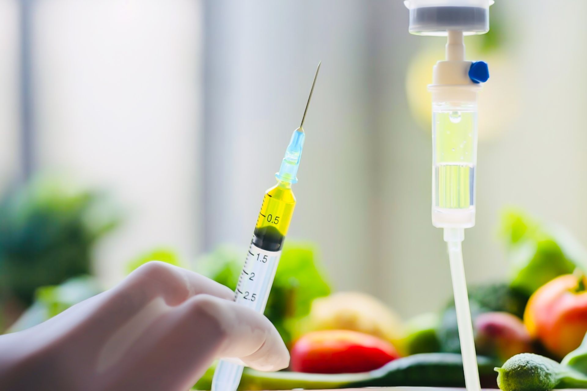 Hand holding a syringe with yellow liquid beside a drip set, with blurred fruit in the background