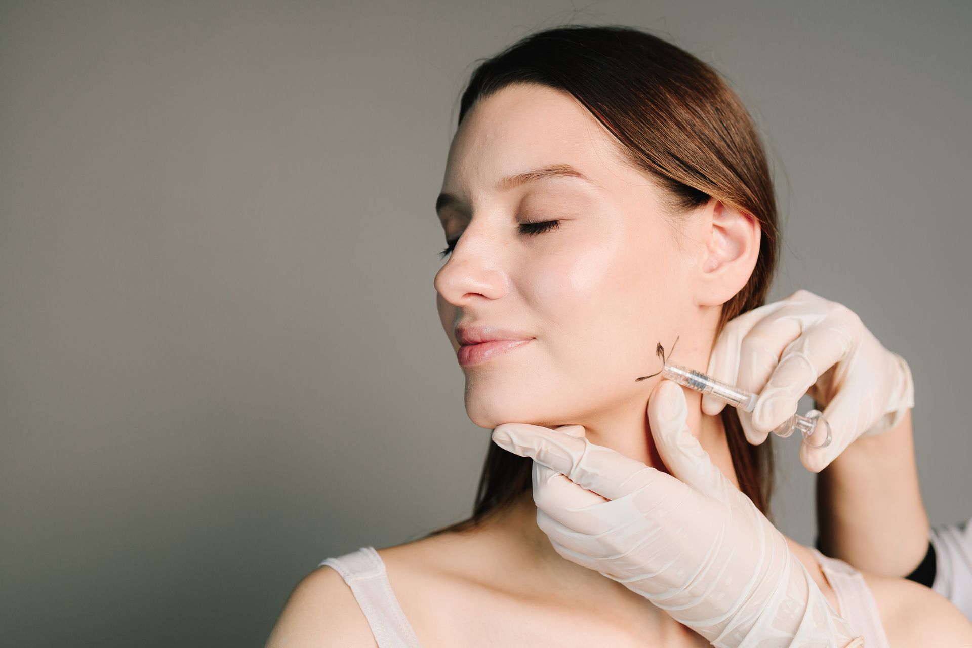 Gloved hands injecting a woman’s jawline with a syringe in a clinical setting