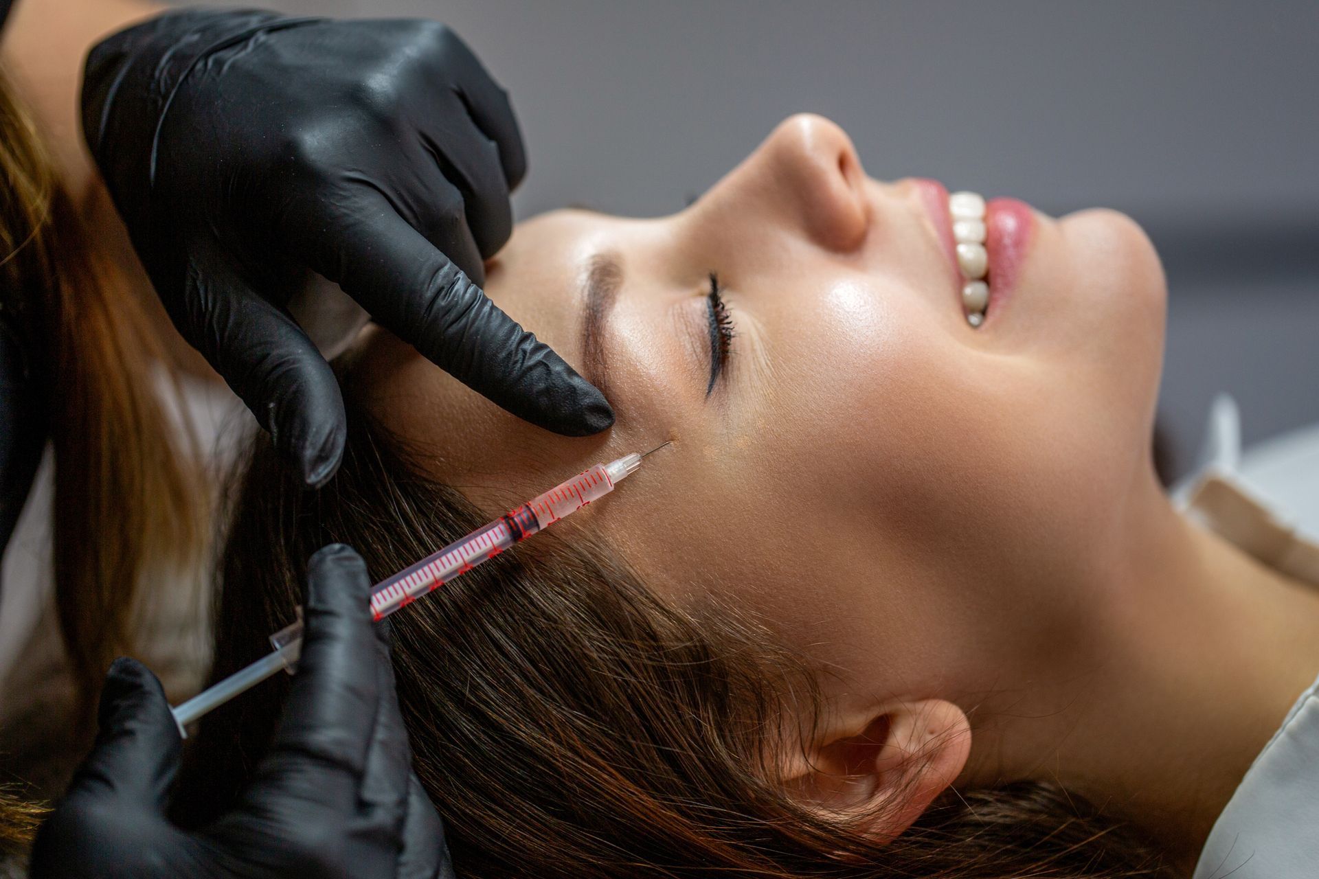 Gloved hands inject facial filler into a smiling patient’s cheek during a cosmetic procedure