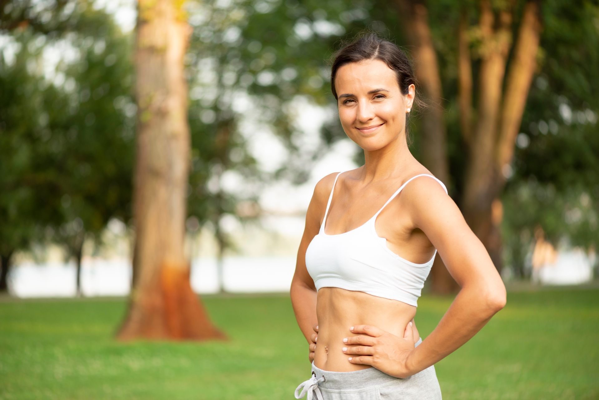 Woman in a white sports bra standing in a park with hands on hips, smiling at the camera.