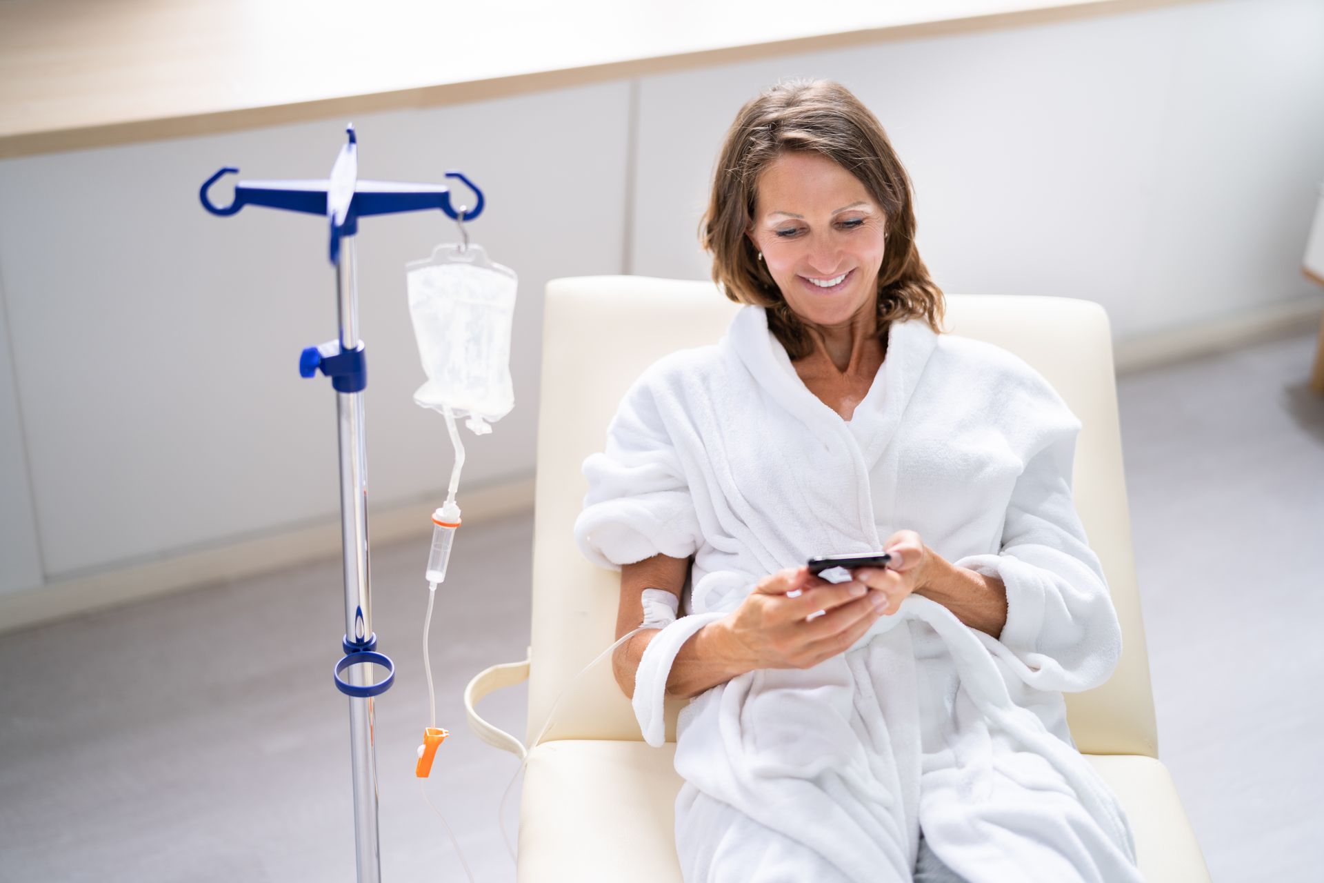 Patient in a white robe sits in a hospital chair, smiling at a phone beside an IV stand.