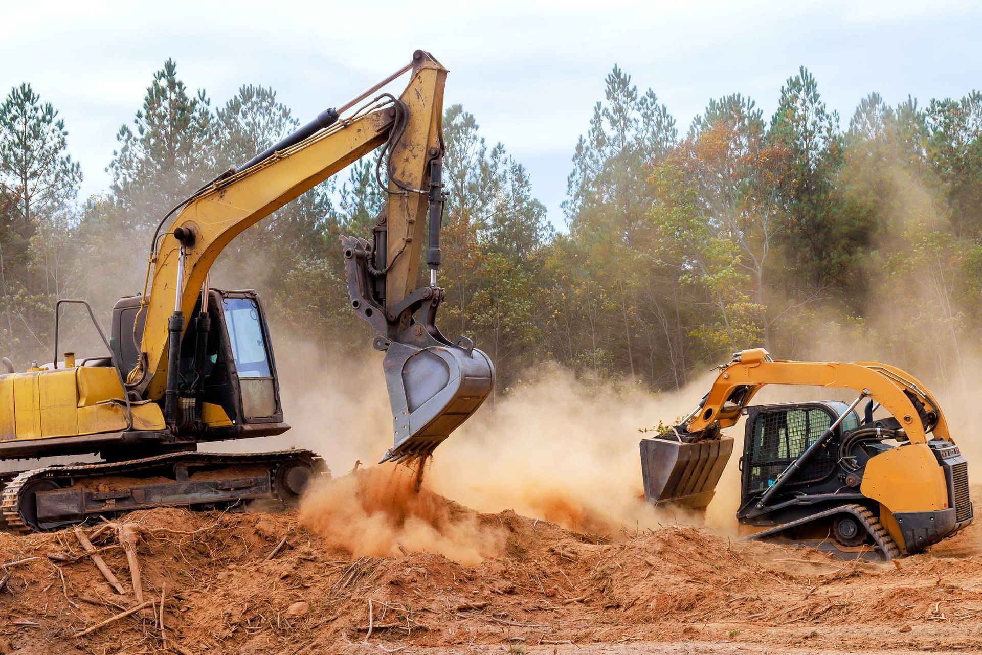 Excavator and skid-steer loader moving dirt at a dusty construction site near trees. Excavator and skid-steer loader moving dirt at a dusty construction site near trees.