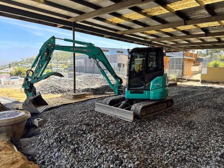 A compact green mini excavator parked under a partially covered structure, positioned on a bed of crushed gravel with construction materials. A compact green mini excavator parked under a partially covered structure, positioned on a bed of crushed gravel with construction materials.
