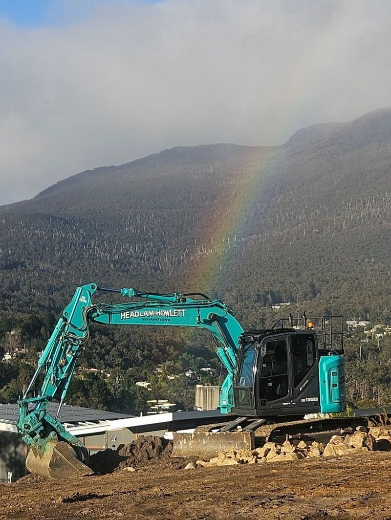 Teal excavator on a construction site with a rainbow arching behind a mountain.