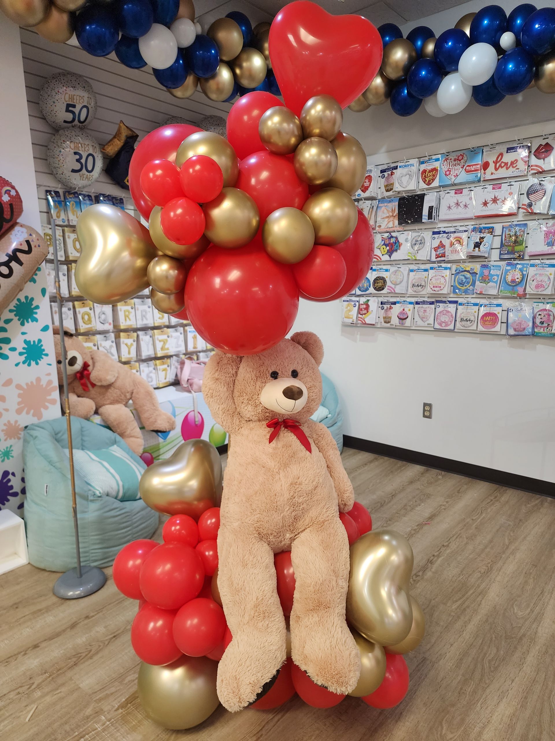 A teddy bear is sitting on top of a bunch of red and gold balloons.