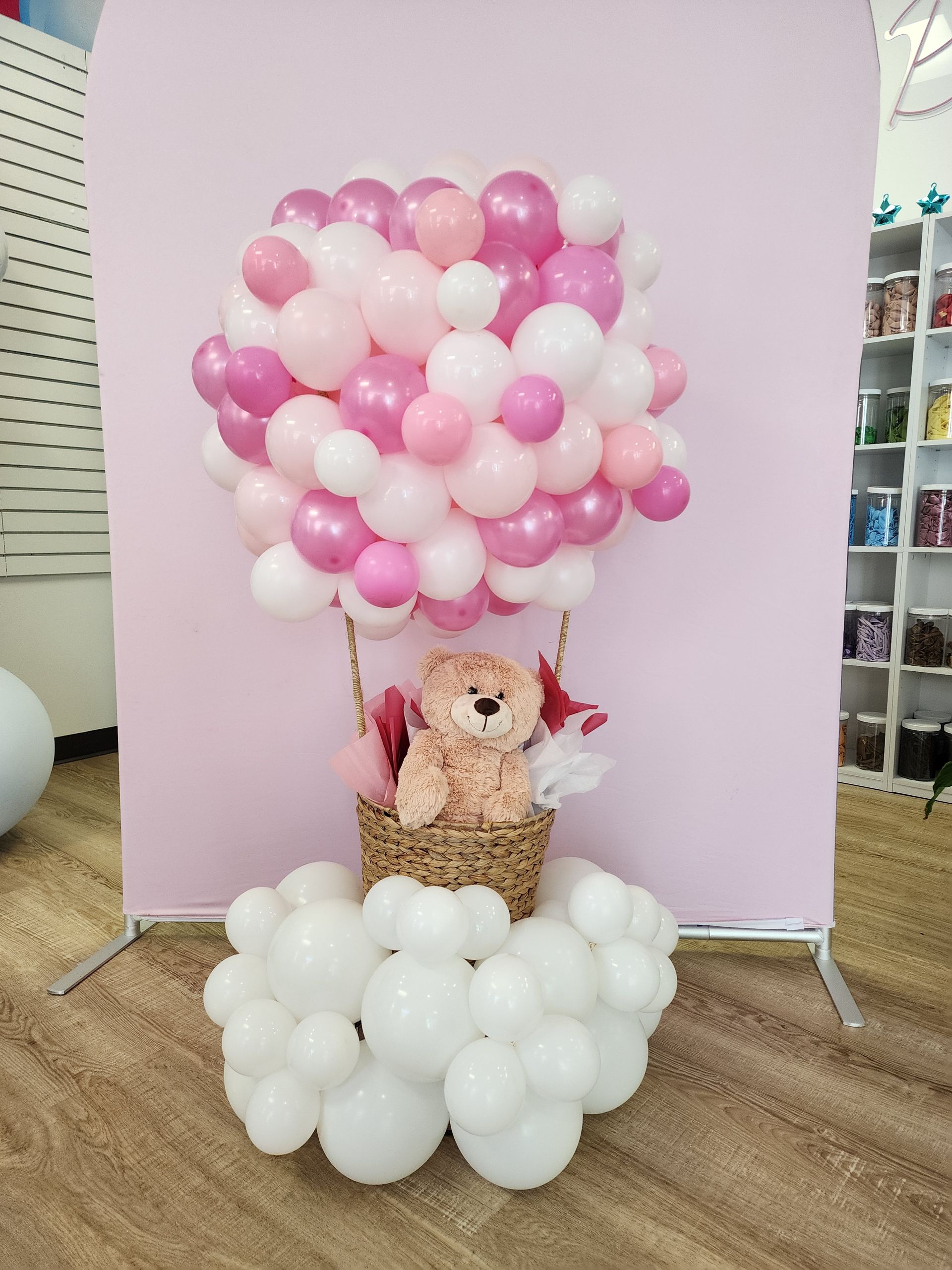 A teddy bear is sitting in a hot air balloon made of pink and white balloons.