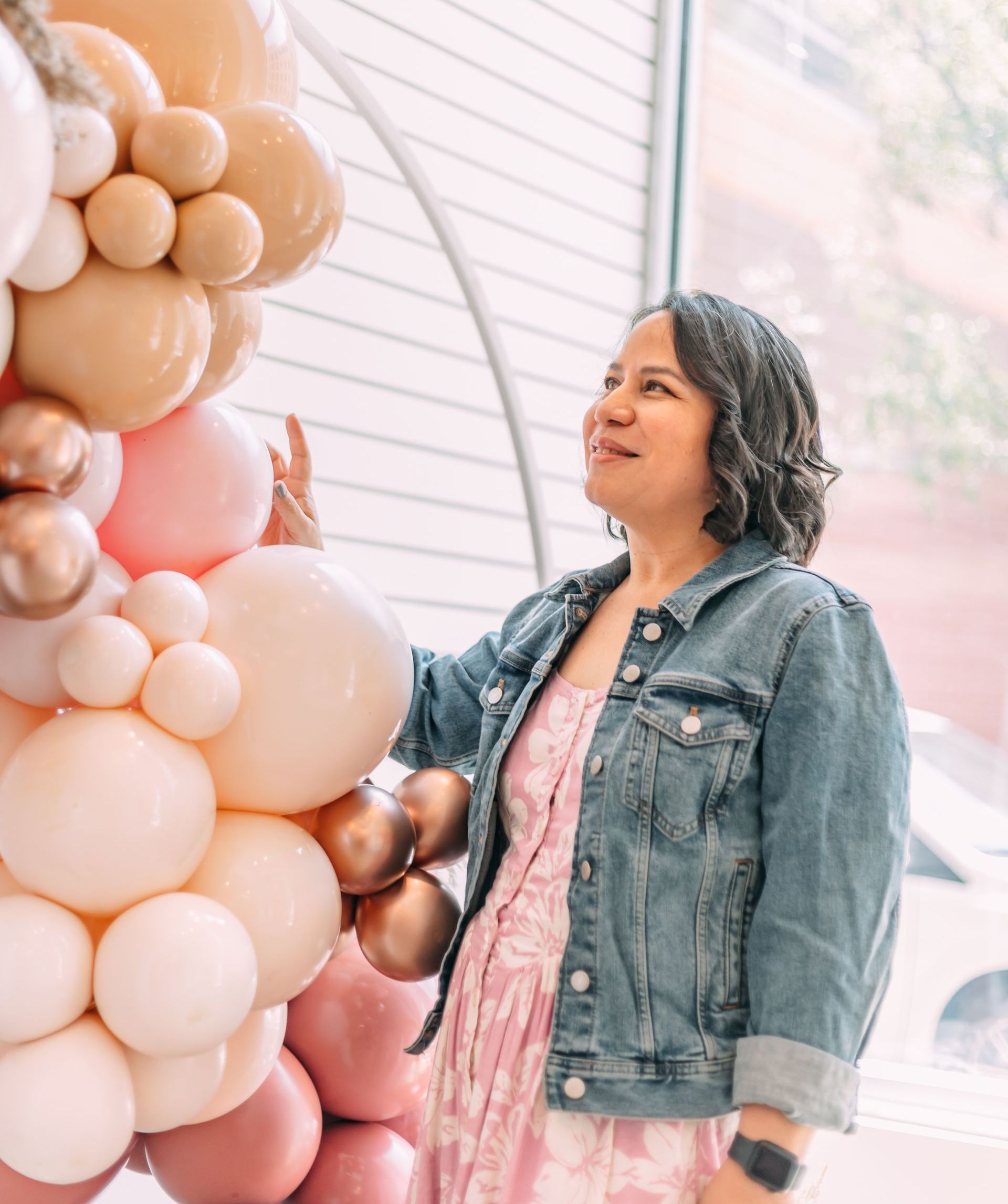A woman in a denim jacket is standing in front of a wall of balloons.