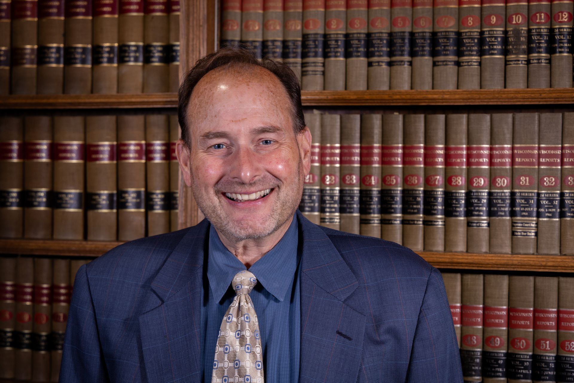 Man smiling in a blue blazer and tie, standing in front of a bookcase filled with books.