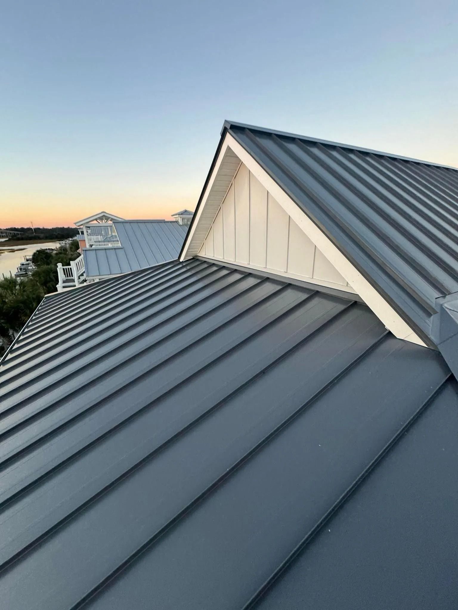 Metal roof at sunset with gray standing-seam panels and white gables overlooking water