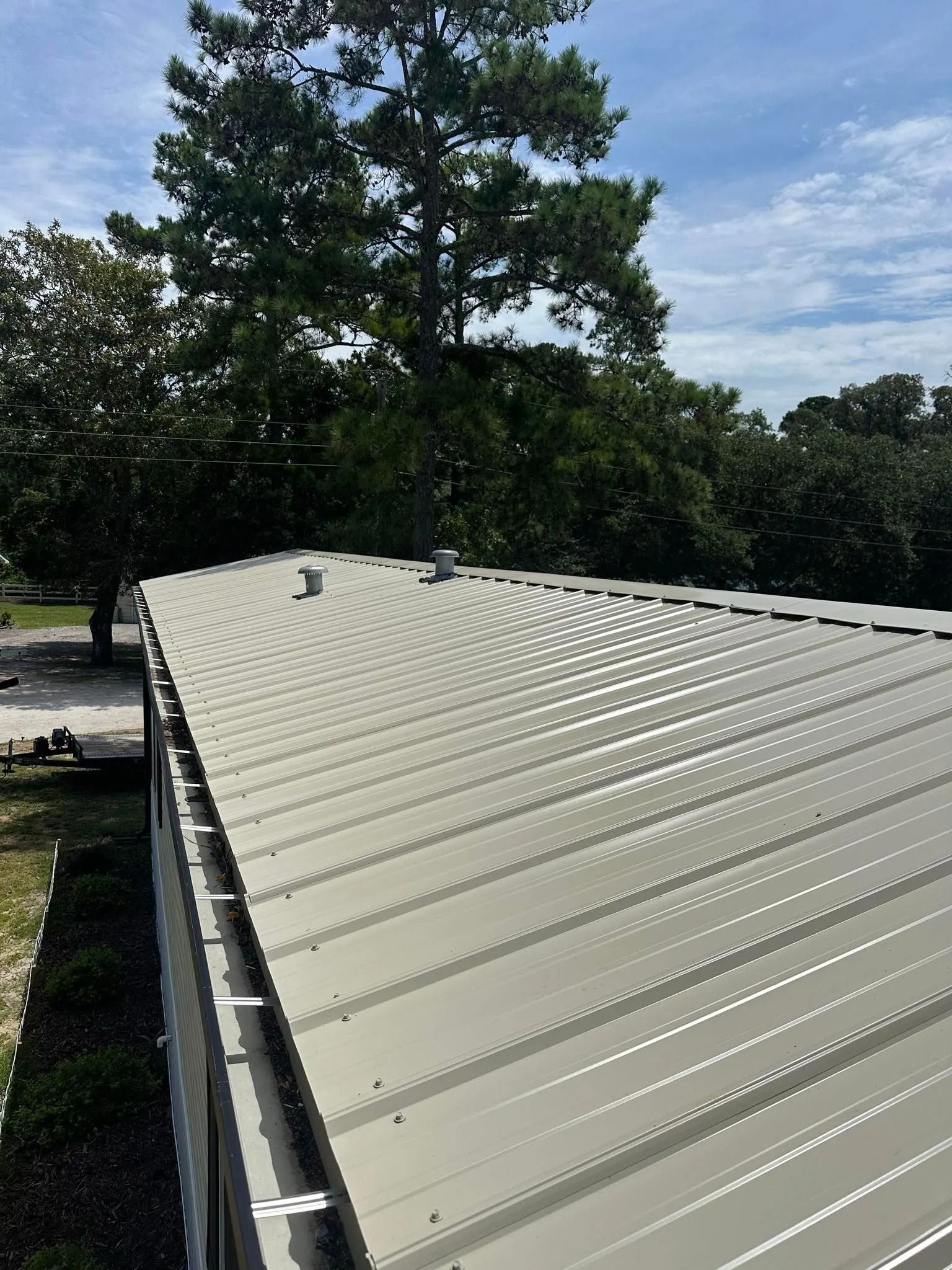 Light beige metal roof with vents, viewed from above beside trees and a cloudy sky