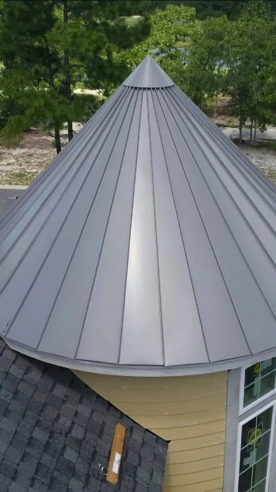 Gray conical metal roof on a turret of a yellow house, surrounded by green trees.