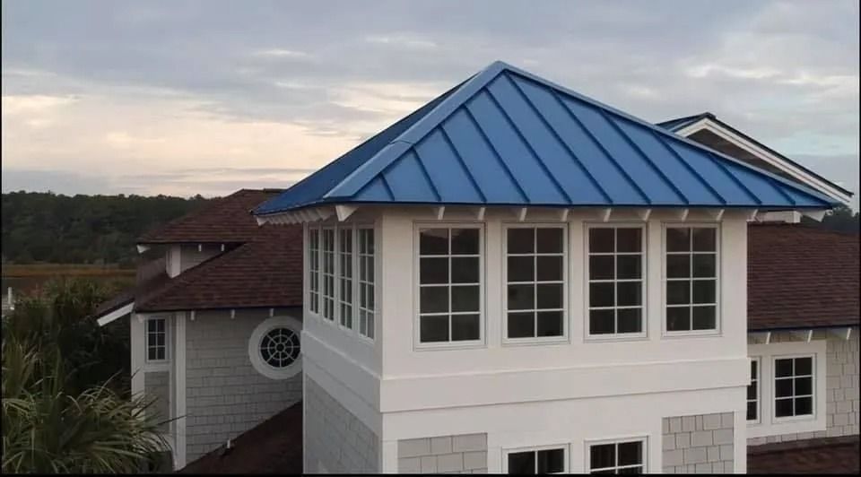 White house with a blue metal roof and dormer windows at dusk under a cloudy sky