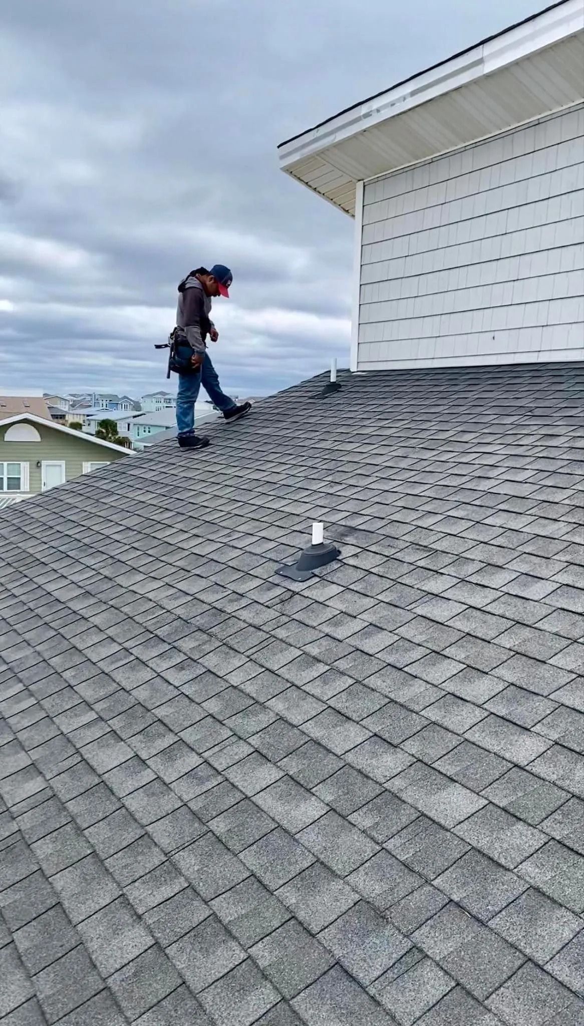 Worker on a steep shingled roof near a white house, carrying equipment under a cloudy sky.