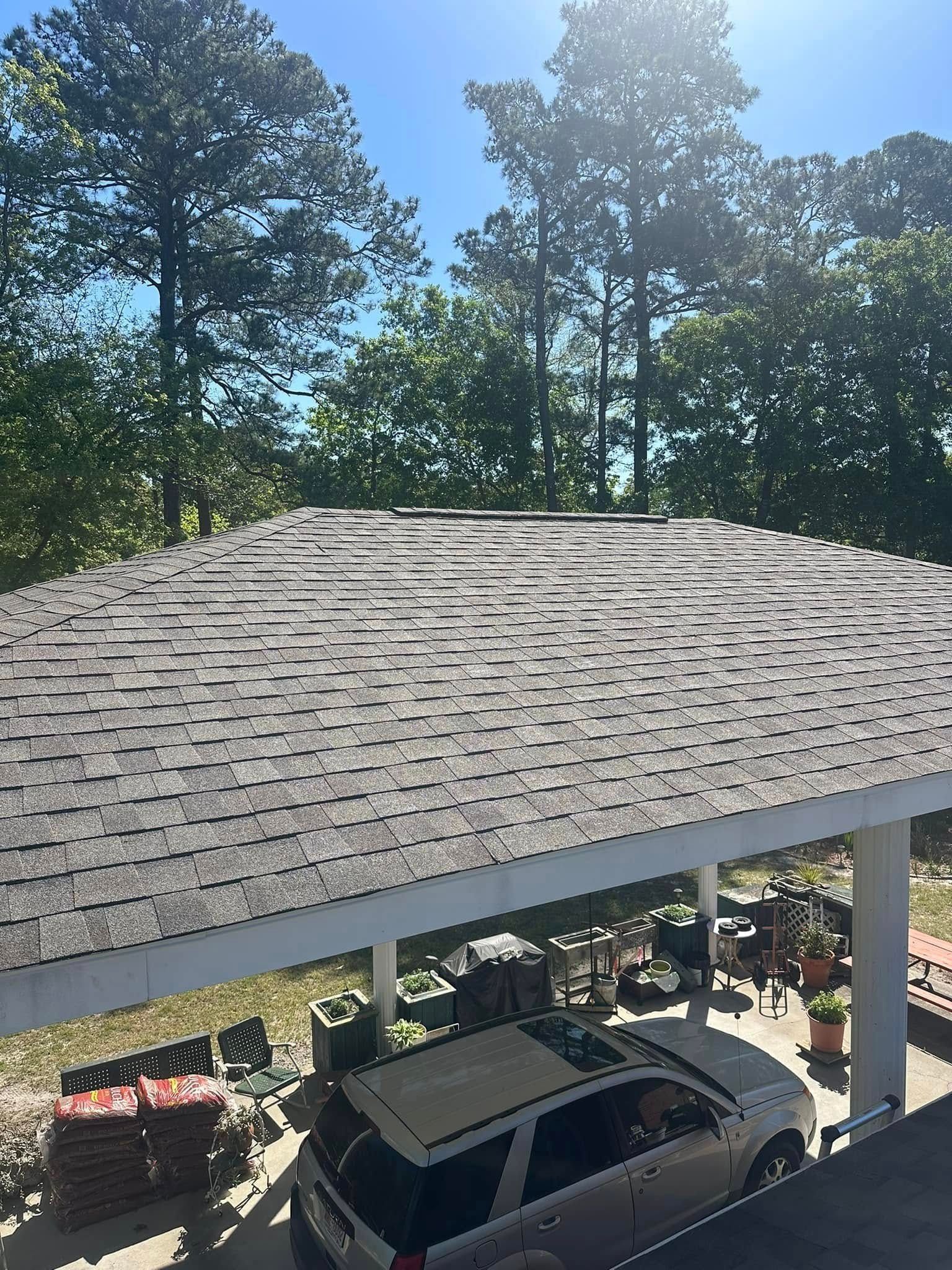 Carport with gray shingled roof and parked car beneath, surrounded by trees in bright sunlight.