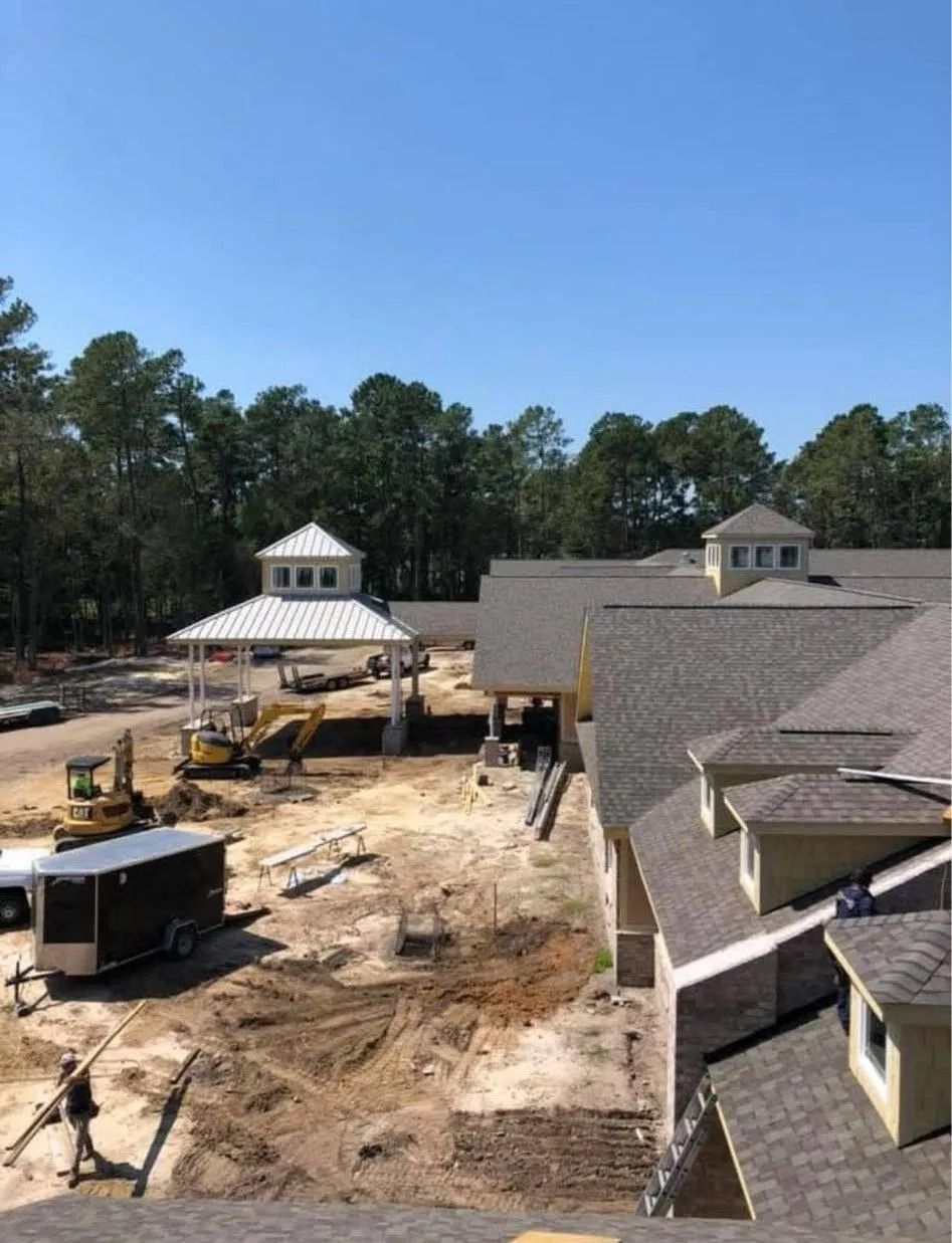 Construction site with dirt lot, yellow excavator, trailers, and rooftops under a clear blue sky