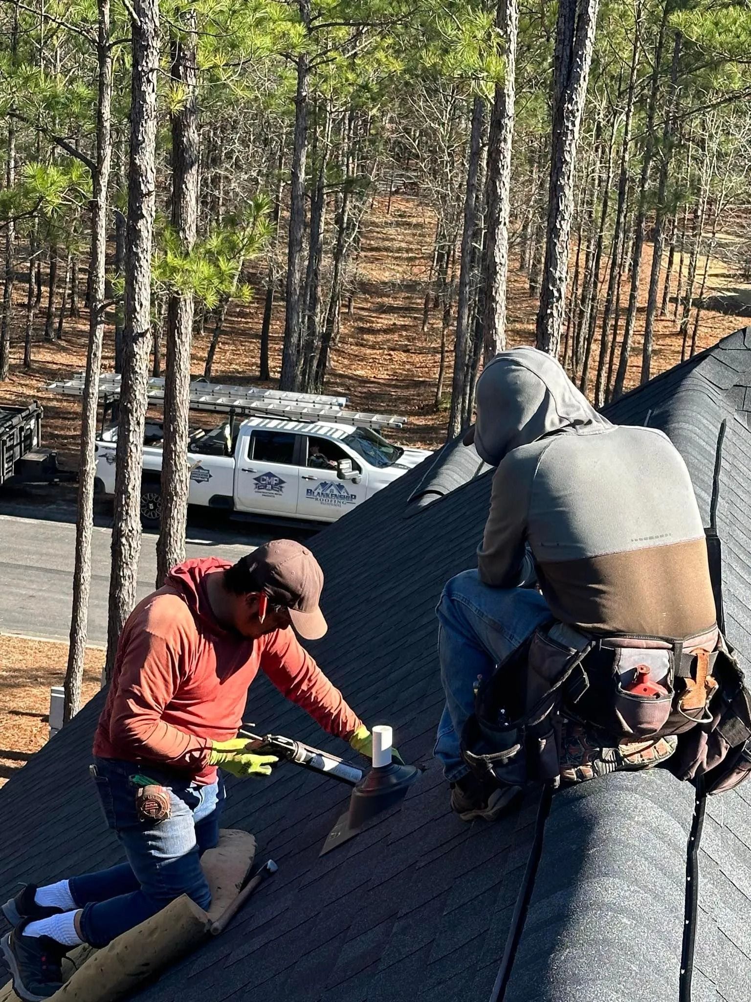 Workers installing roof shingles on a house, with one crouched and another kneeling on the roof.