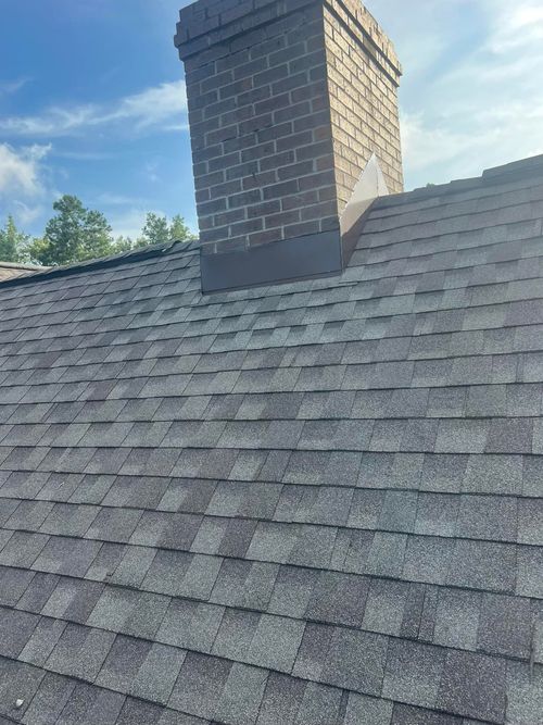 Shingled roof with a brick chimney against a blue sky and trees