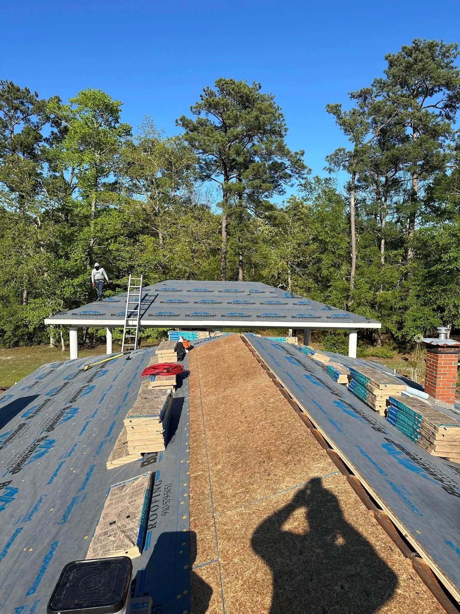 Workers roofing a house with blue tarp, seen from the roof ridge on a sunny wooded site.
