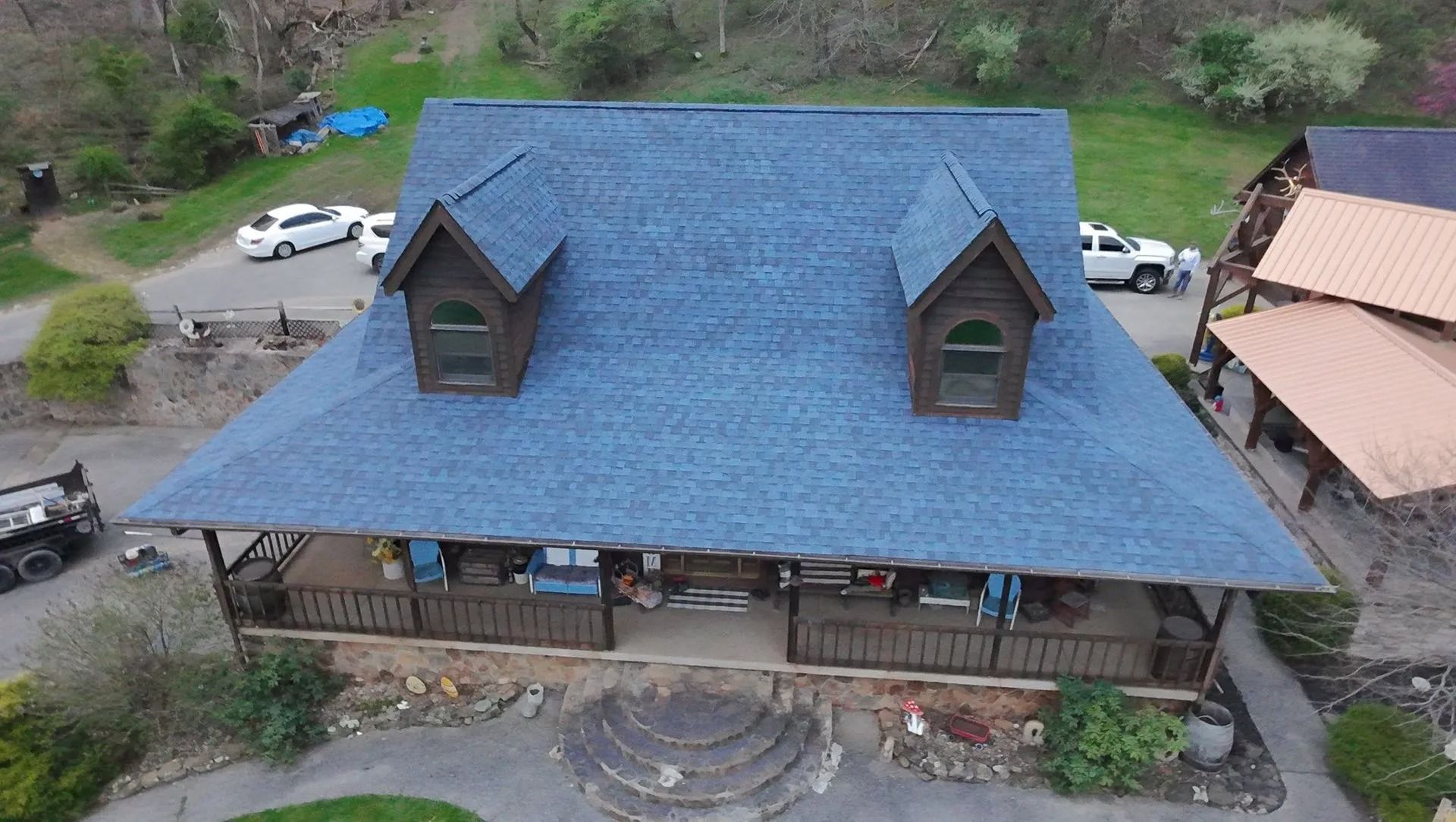 Aerial view of a large rustic house with a blue roof and front porch, surrounded by trees and parked cars.