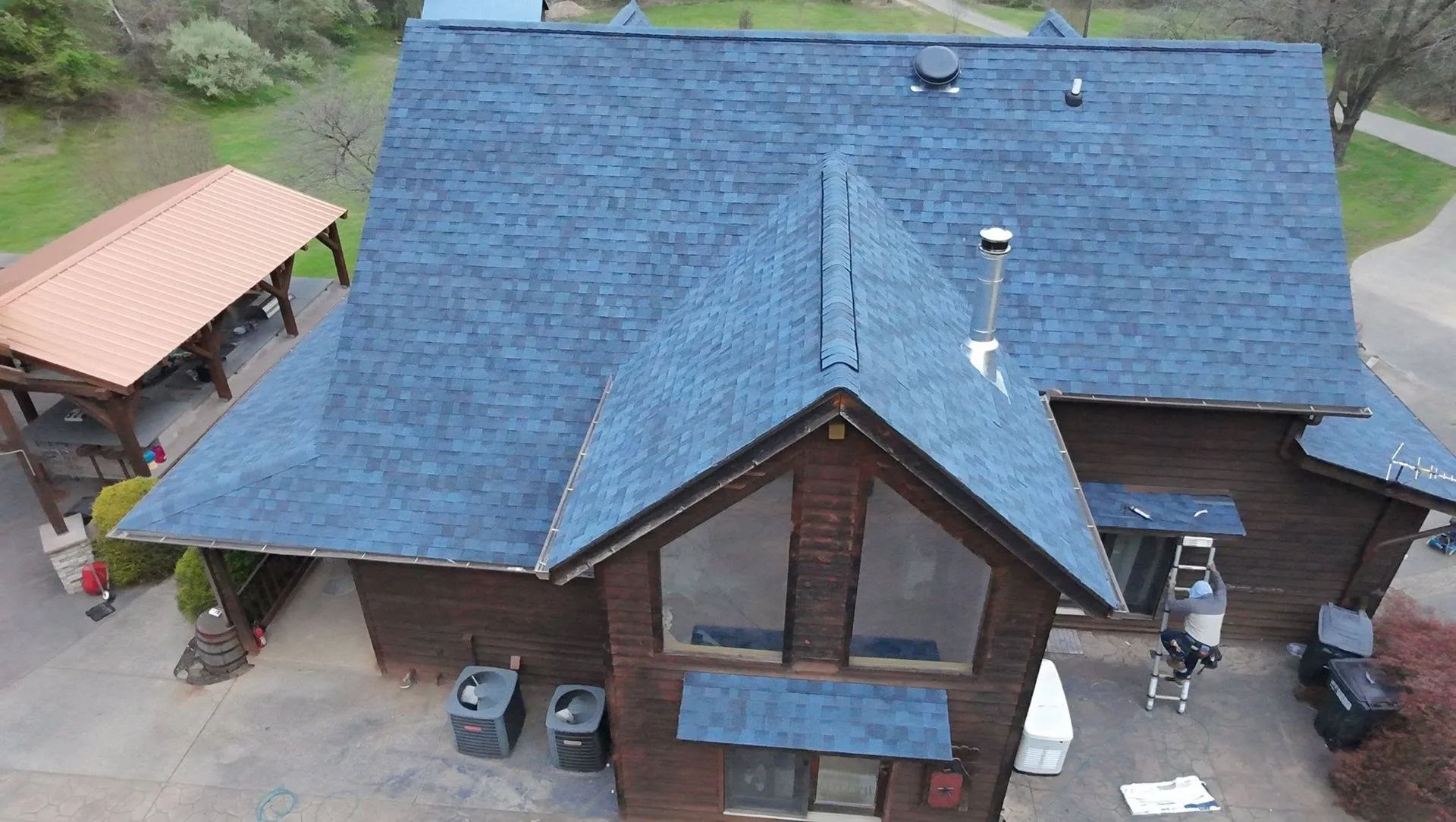 Aerial view of a brown house with a blue roof and a small patio, surrounded by trees and greenery