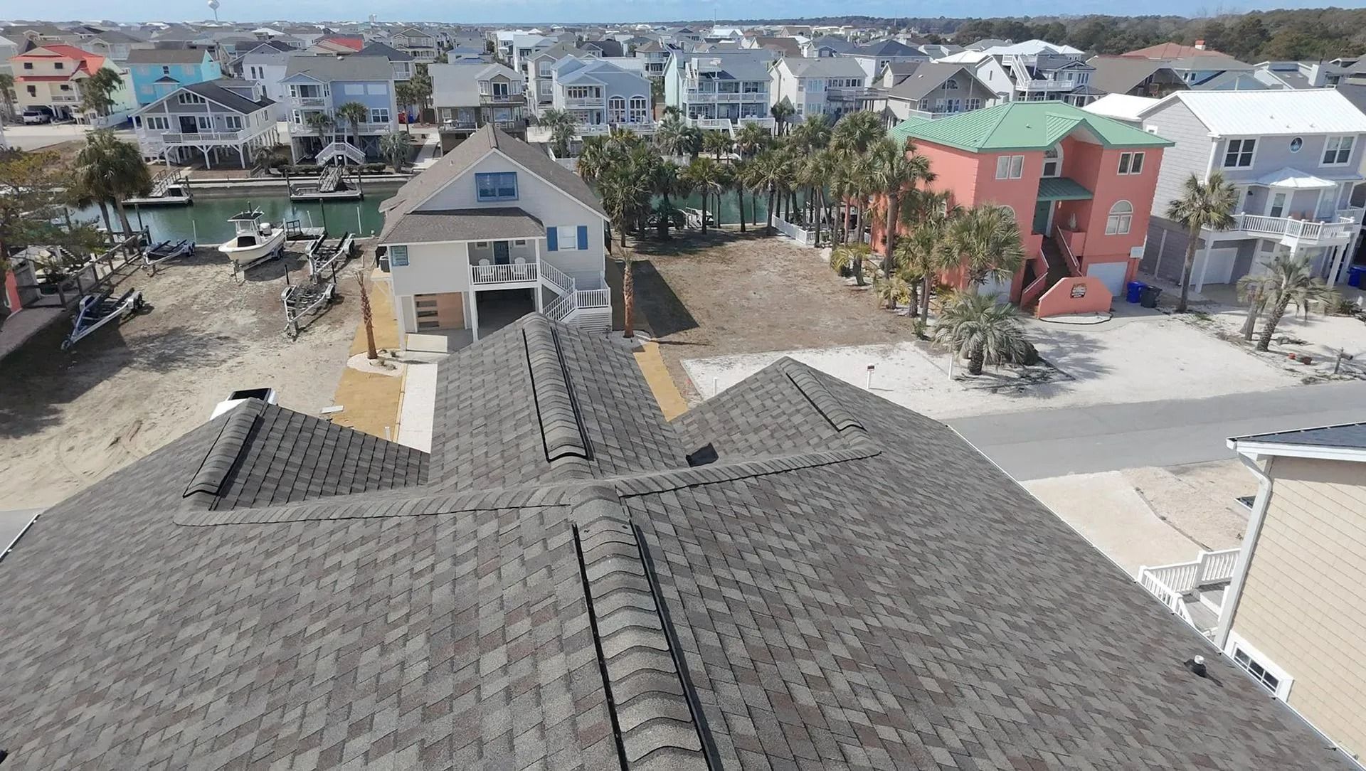 Rooftop view of a coastal neighborhood with colorful houses, palm trees, and parked cars under a clear sky.