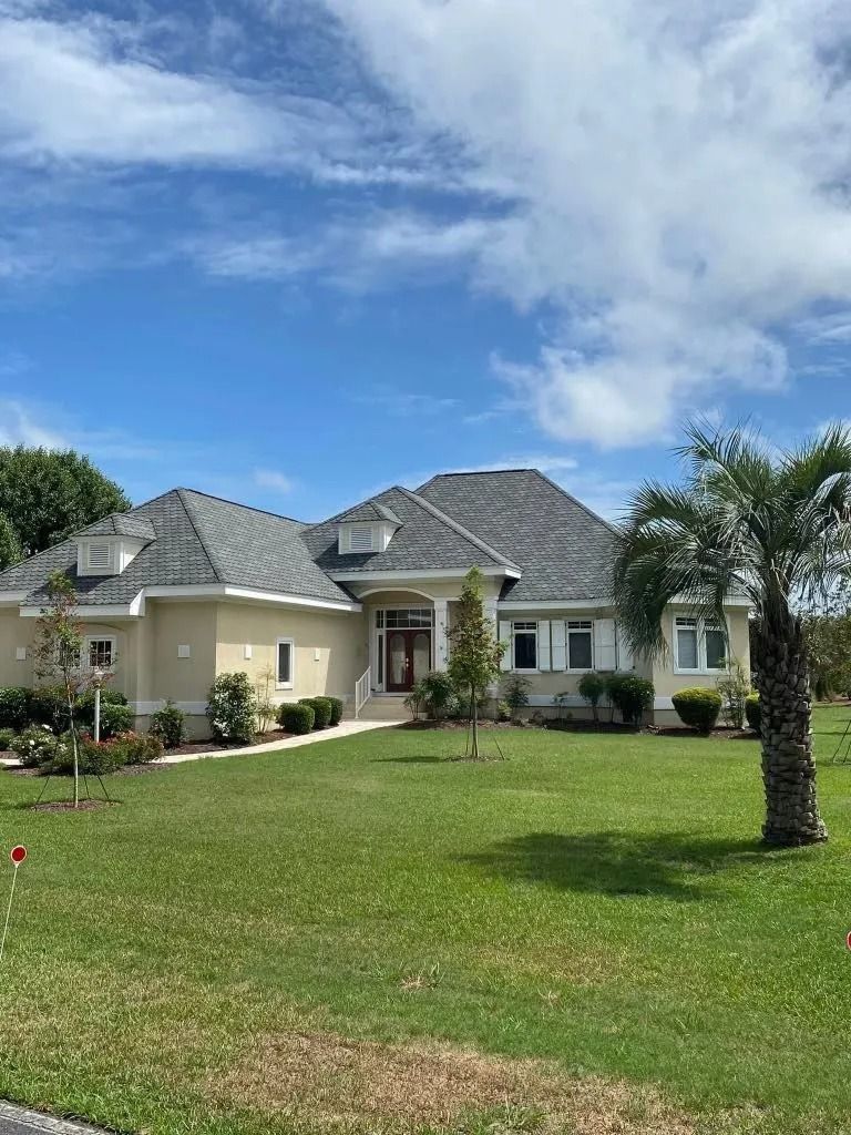 Single-story cream house with gray roof and palm tree in a green lawn under a blue sky