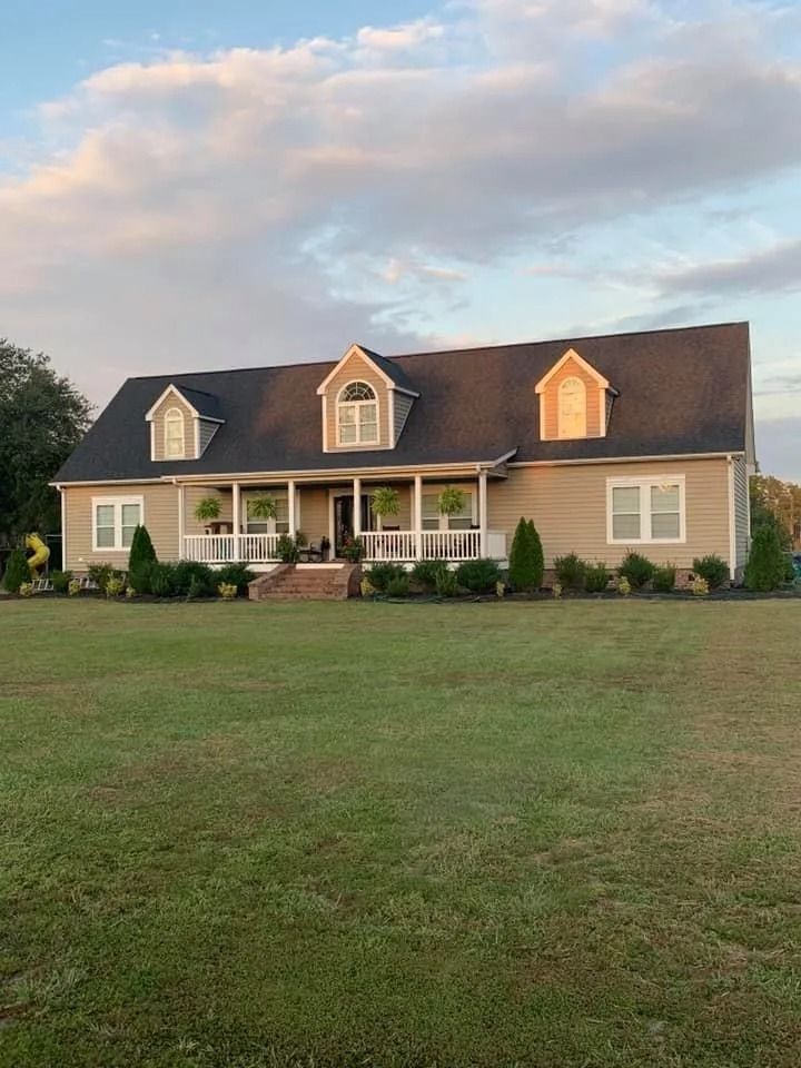 Single-story beige house with a dark roof and front porch, set on a large green lawn at sunset