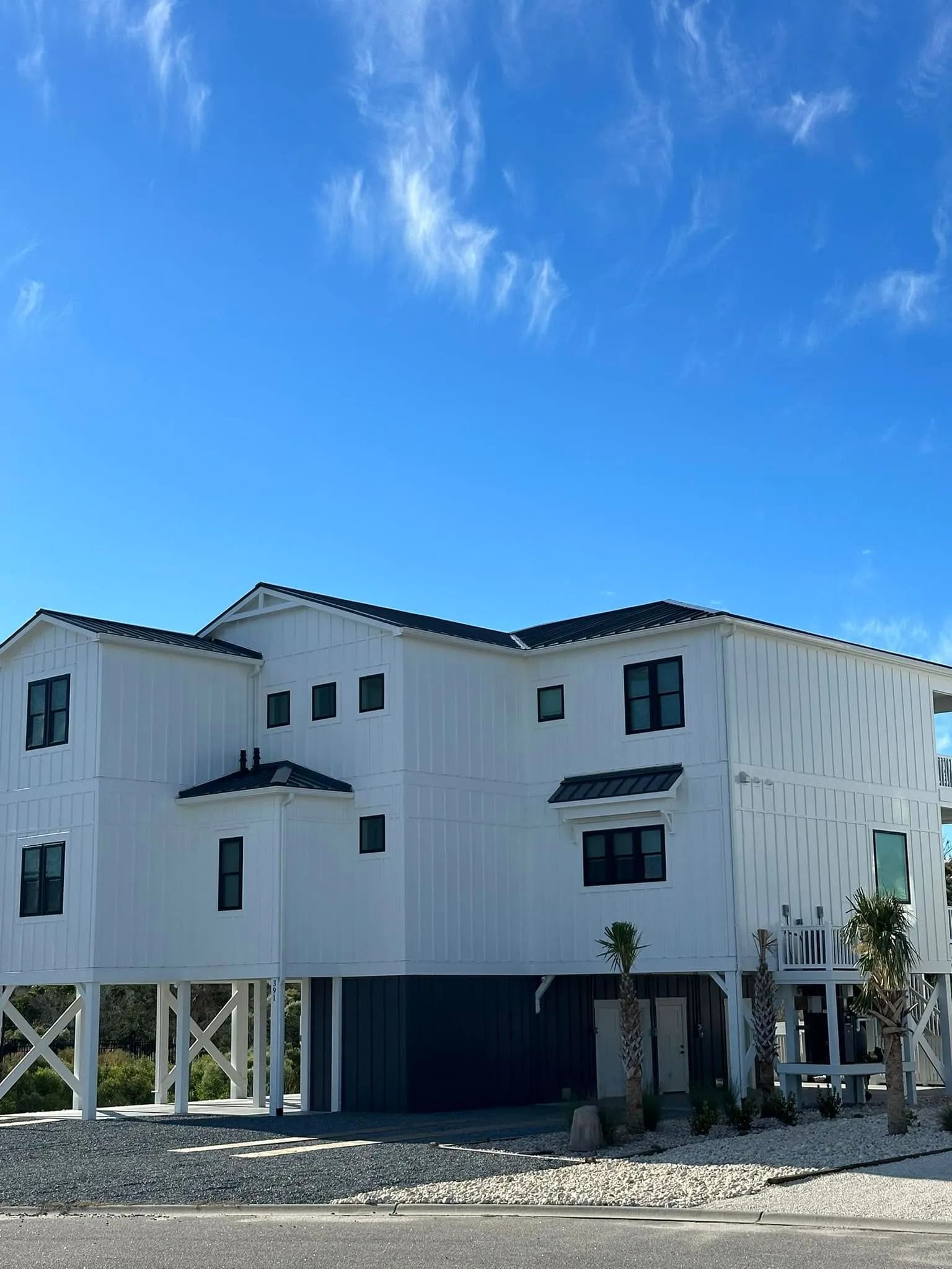 White multi-story house with dark roof and windows on stilts under a clear blue sky