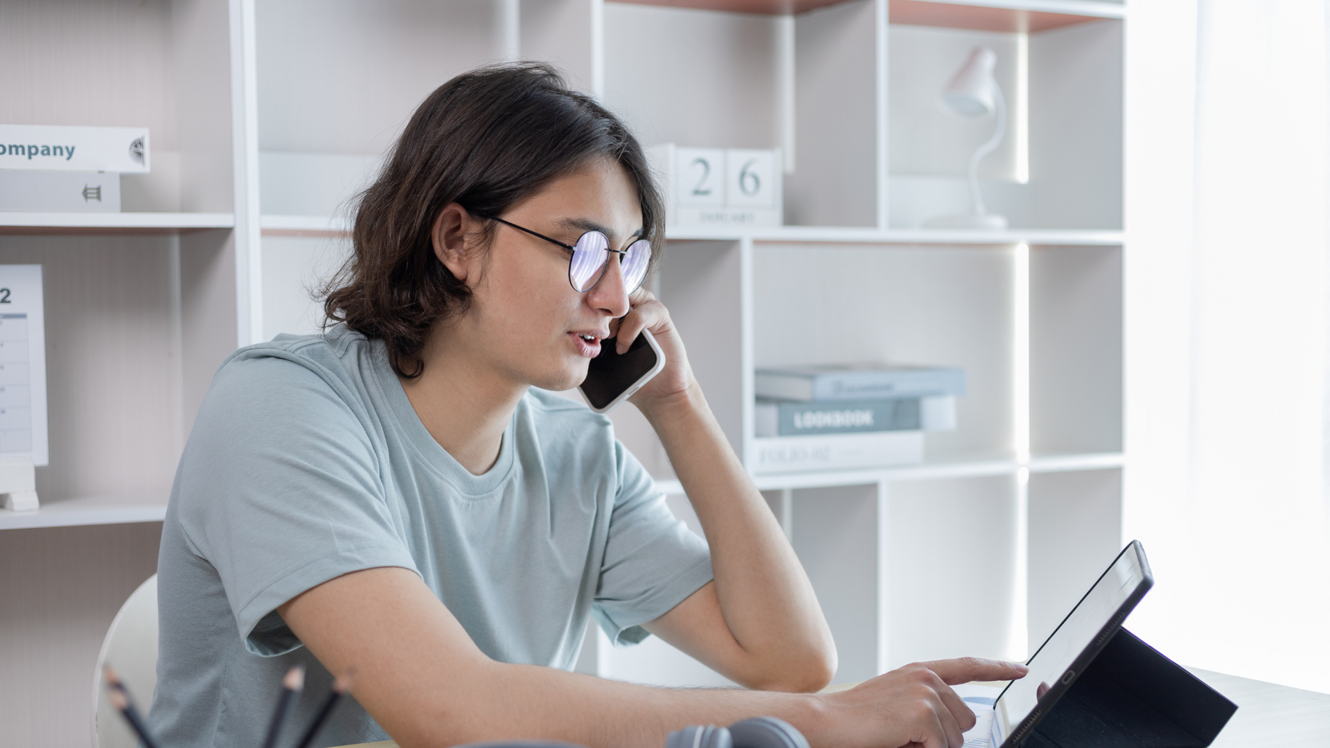Man with glasses on the phone, looking at a tablet in a bright home office.