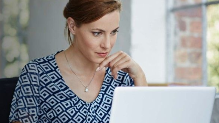 Woman with red hair, looking intently at a laptop. She is wearing a blue patterned shirt.