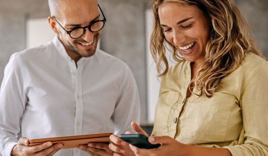 Man with glasses and woman smiling, looking at a phone. Man holds a tablet.