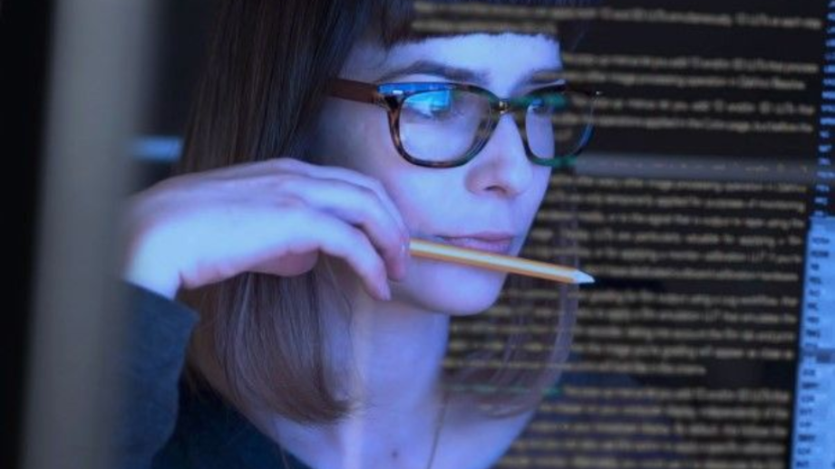 Woman with glasses, holding a pencil, looking at a glowing computer screen. Indoor setting, possibly a tech office.