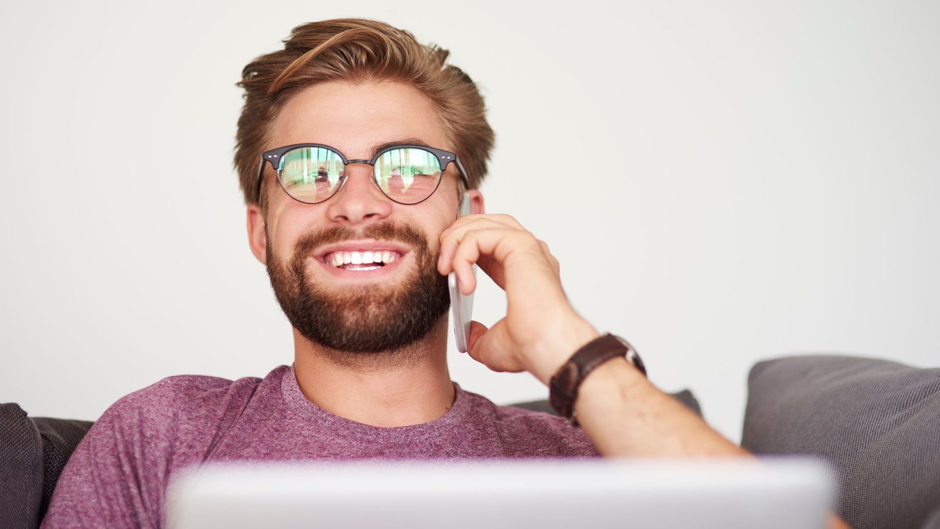 A man with a beard is sitting on a couch talking on a cell phone.