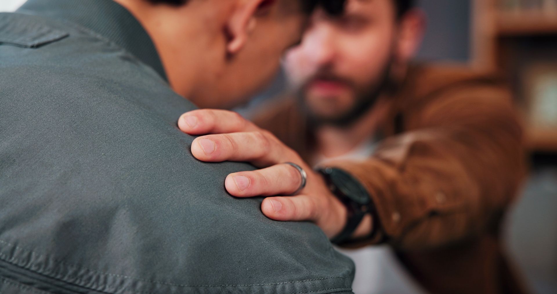 Hand on a person’s shoulder in a blurred indoor setting, suggesting support or comfort