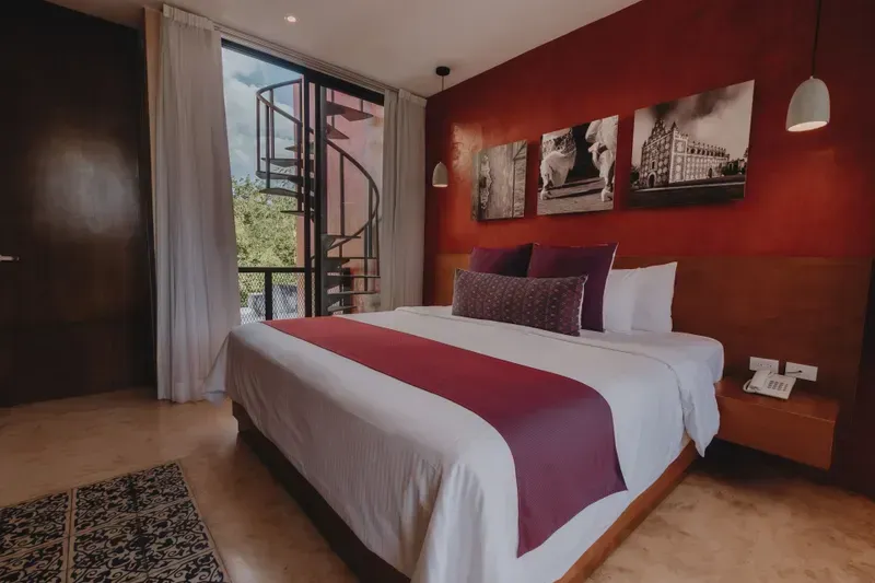 Modern hotel room with a large bed, red accent wall, and balcony door letting in daylight