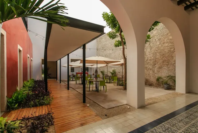 Modern courtyard with arches, terracotta tiles, tables, chairs, and white umbrellas beside a coral wall