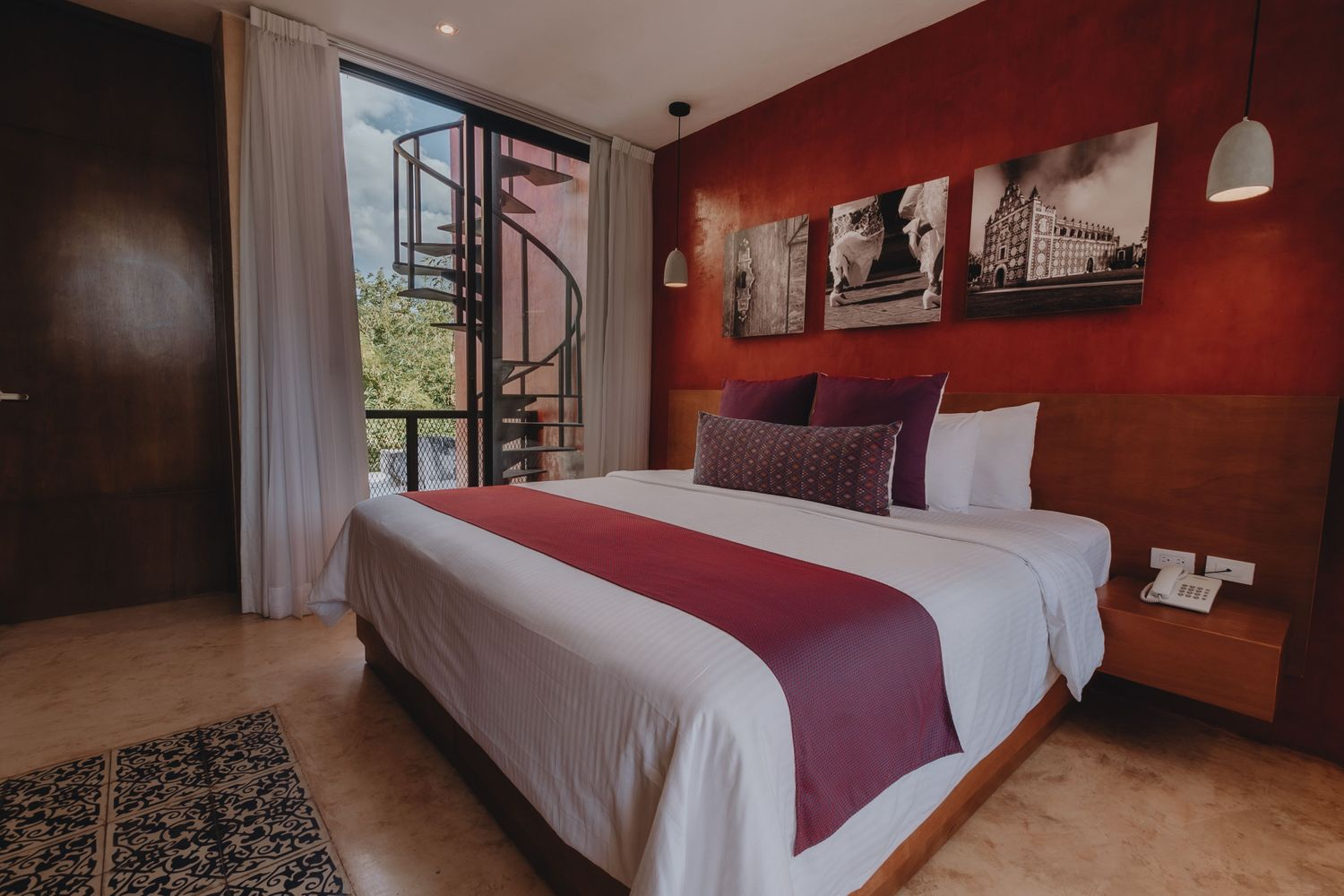 Modern bedroom with red accent wall, large bed, wooden furnishings, and balcony doors open to greenery