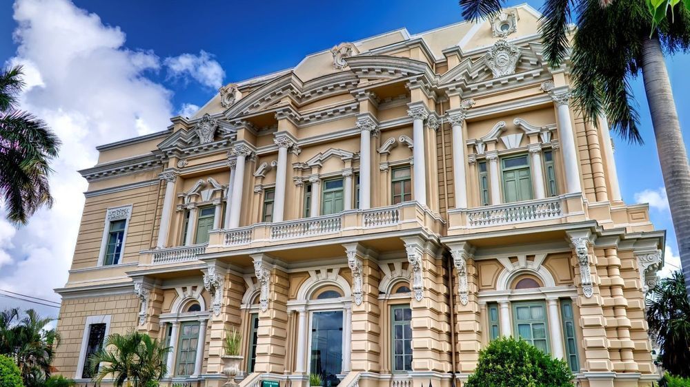 Ornate historic mansion with cream stone facade, arched windows, and blue sky palm trees above.