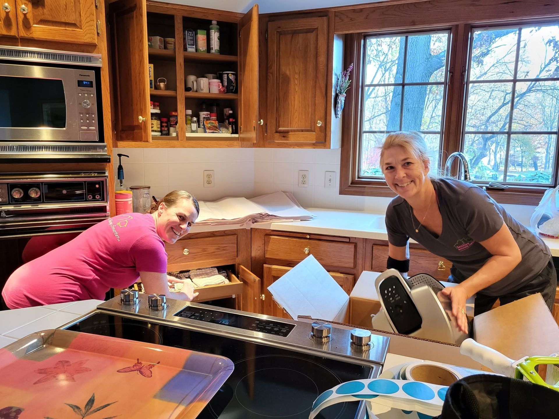 Two people unpacking boxes in a kitchen with wooden cabinets and a window