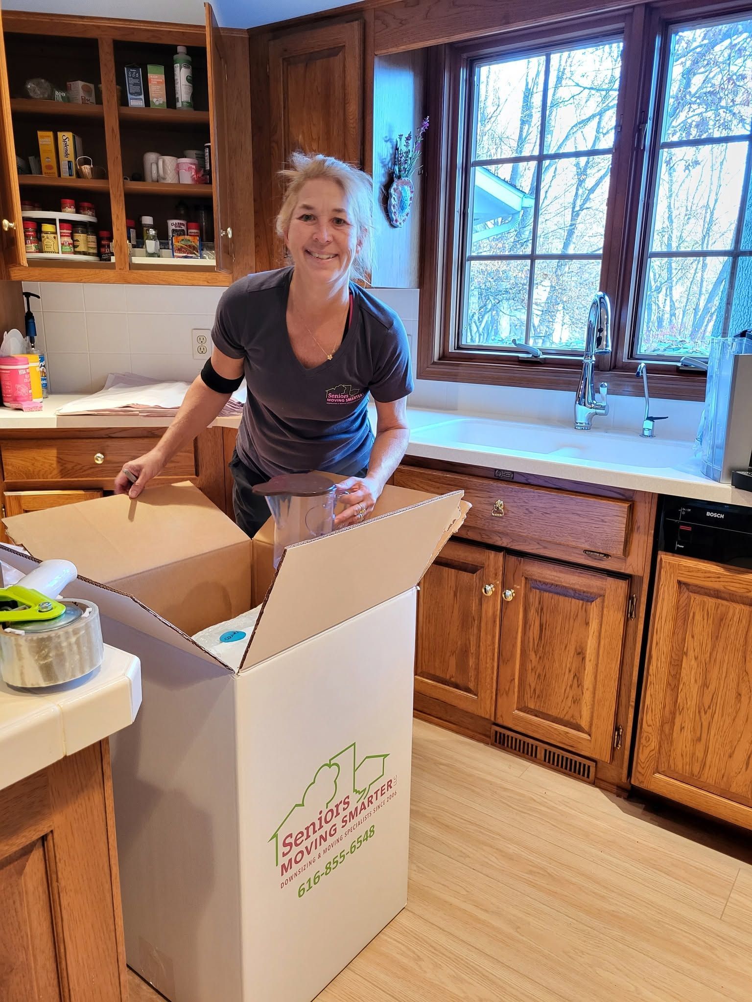 Smiling person in a kitchen, leaning into an open box by the counter near a sunny window.
