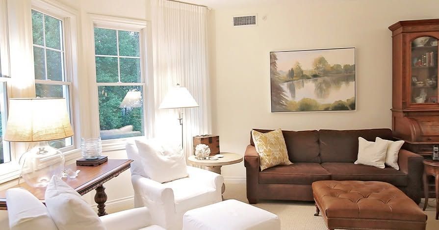 Bright living room with brown sofa, white chairs, large windows, and lamps.