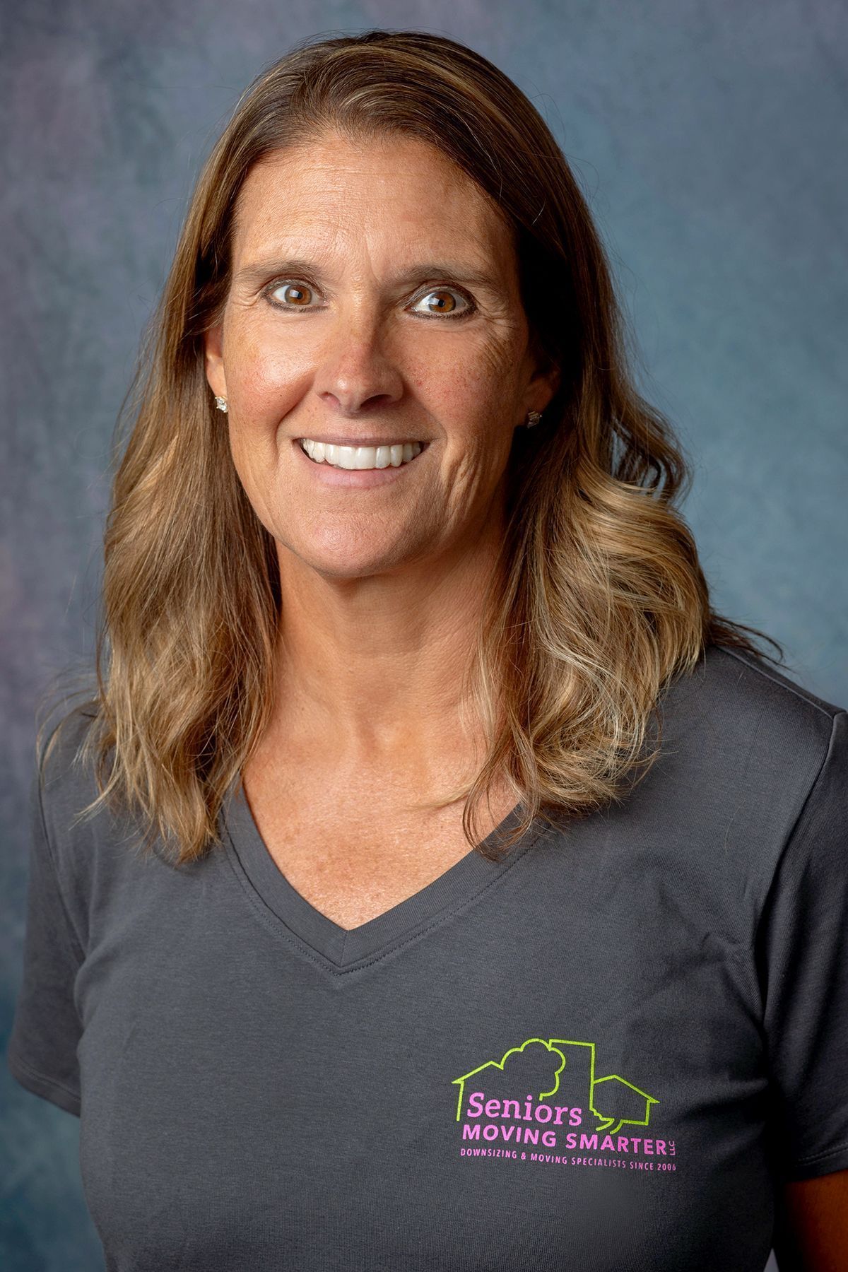 Portrait of a smiling person in a gray V-neck shirt against a blue-gray studio background.
