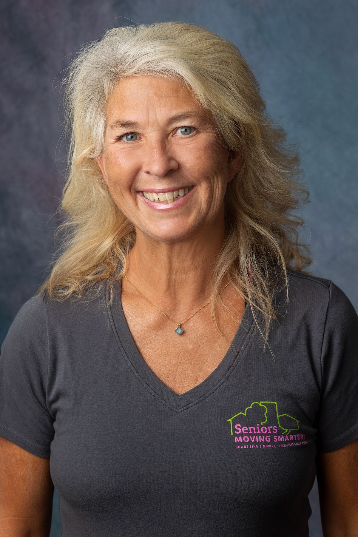 Smiling woman with long blonde hair wearing a dark gray T-shirt against a gray studio backdrop