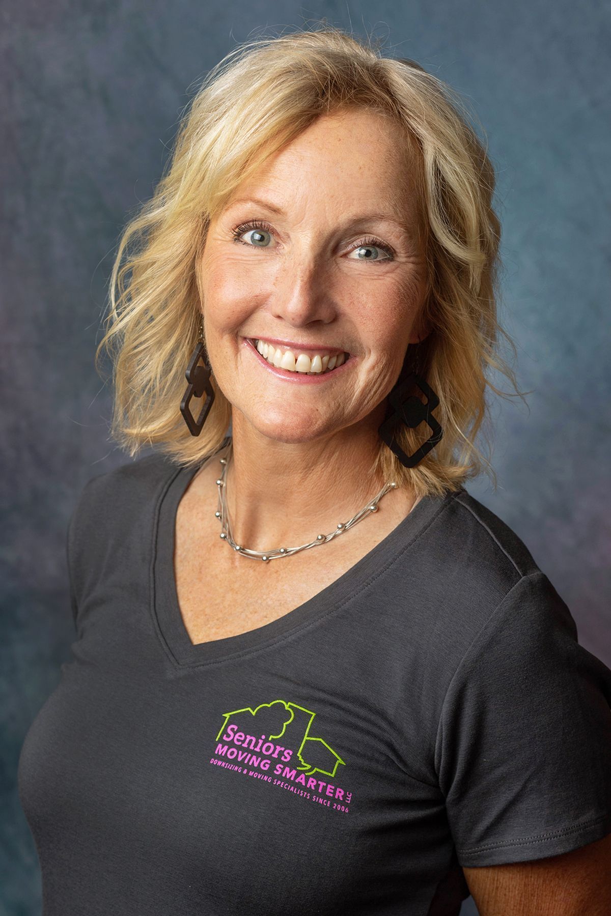 Smiling woman in a dark gray T-shirt with a logo, posed against a gray studio backdrop