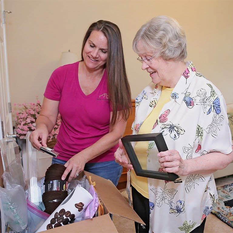Two women unpack gifts, one holding a framed photo, beside open boxes and wrapped items.