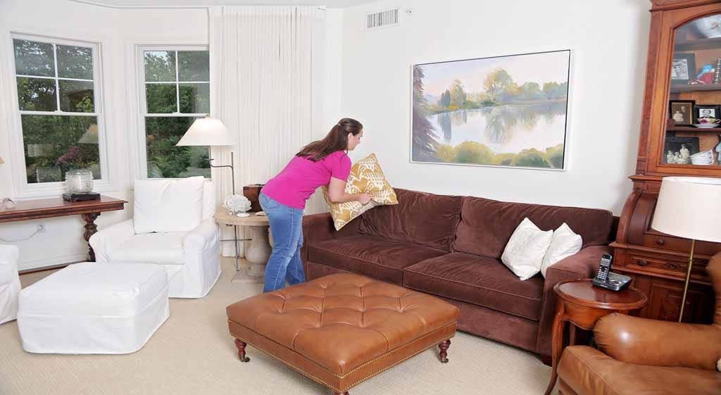 Woman arranging pillows on a brown sofa in a bright living room with white chairs and a large wall painting